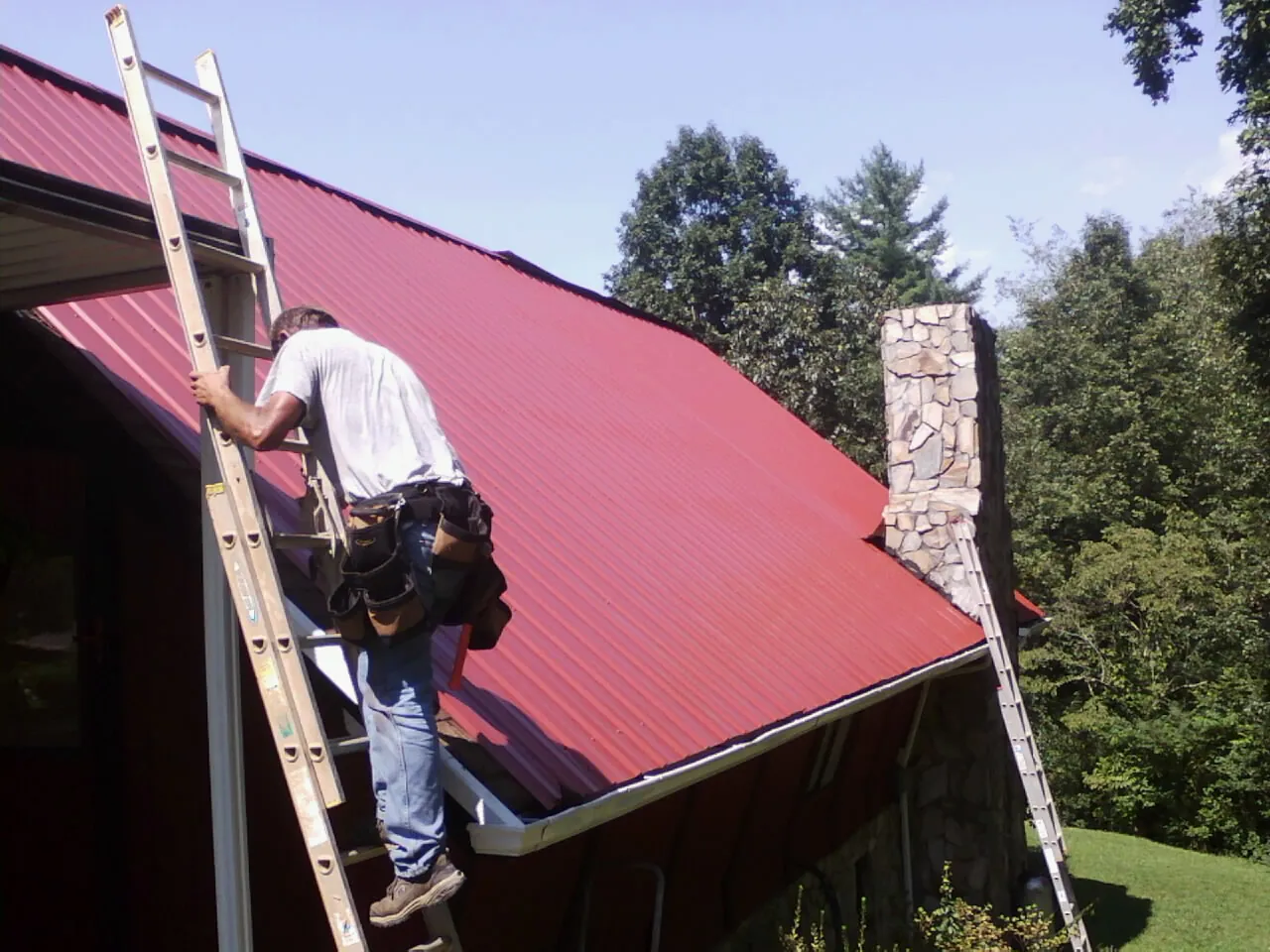 Roofer on a ladder beside a red metal roof and stone chimney — Soffit & Fascia Repair by Roofing Brothers in North Walpole